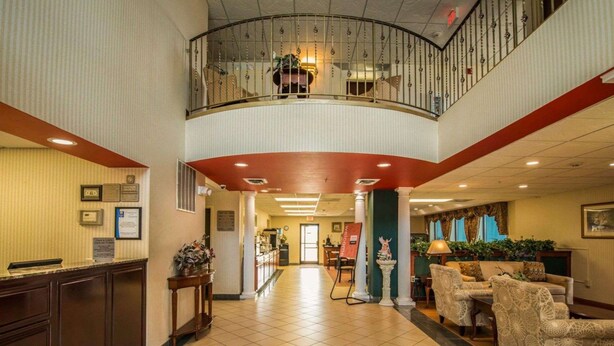 Um hall de hotel com balcão de recepção, área de estar com cadeiras, escadaria e uma fonte decorativa.