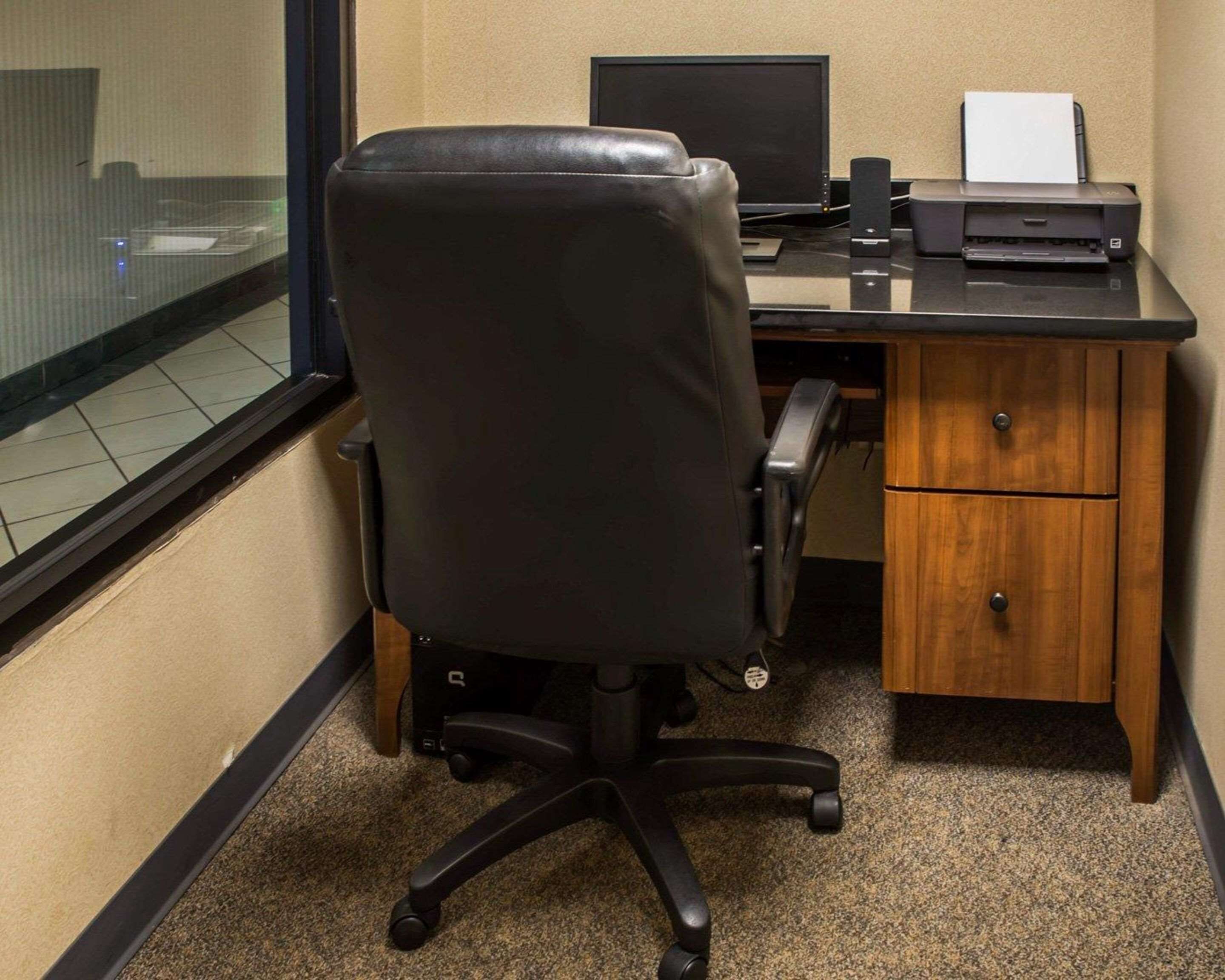 A corner office with a black leather chair, a desktop computer, a printer, and a wooden desk with two drawers.