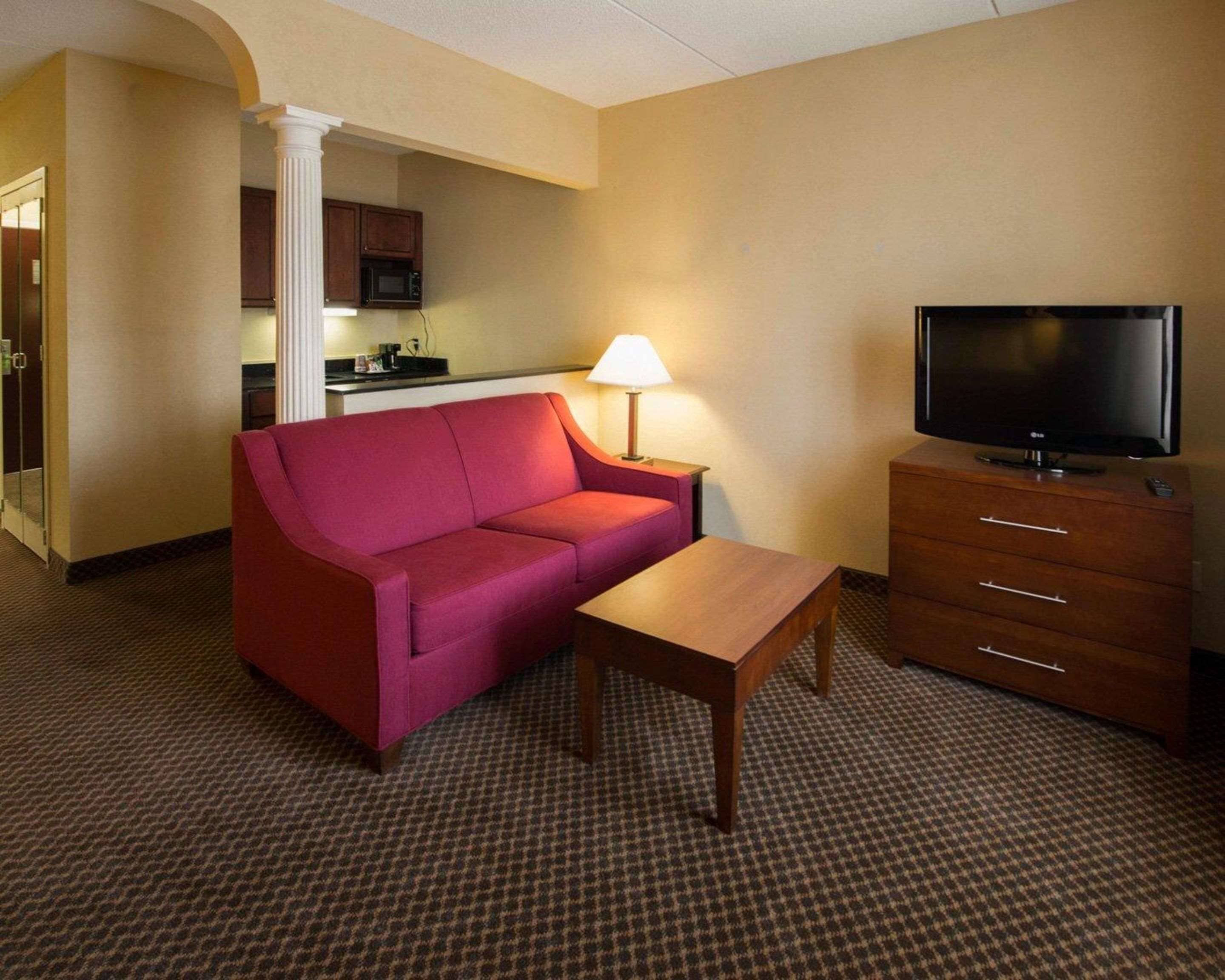 A hotel room with a red sofa, a wooden table, a TV on a wooden cabinet, a lamp, and a kitchenette in the background.