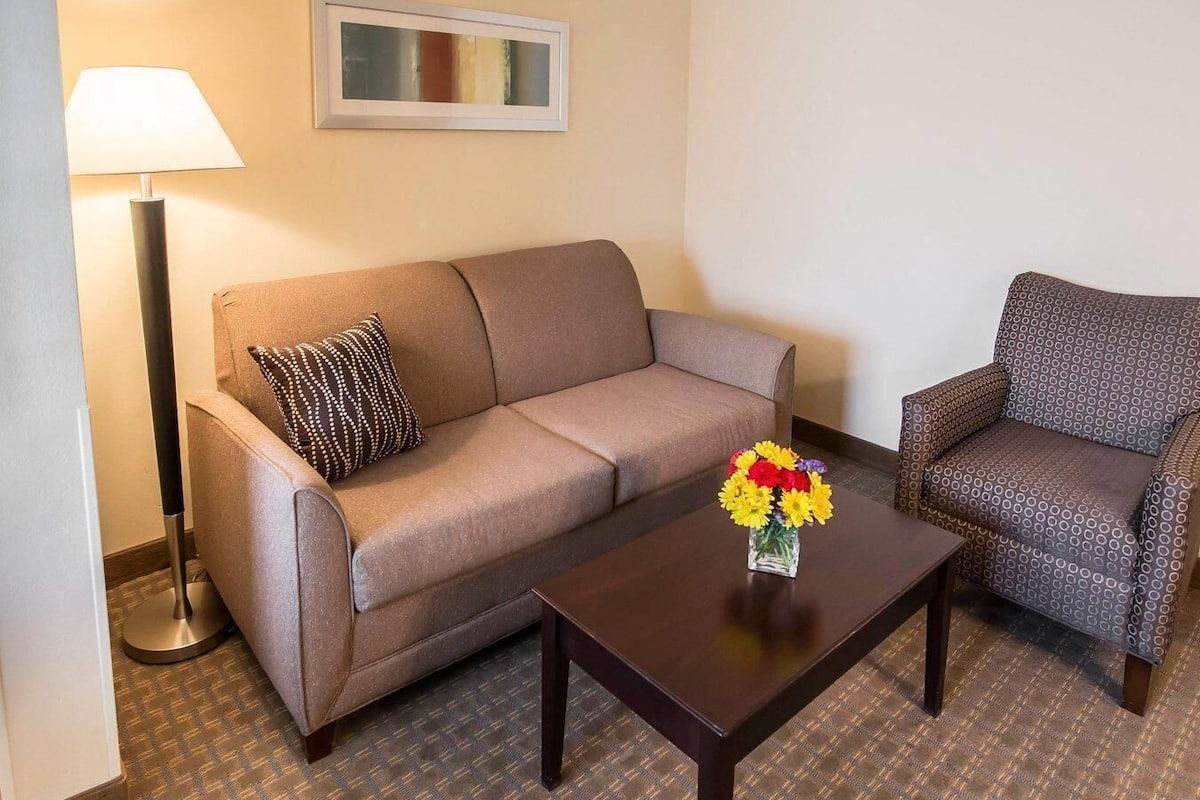 A hotel room with a sofa, armchair, a small table with a vase of flowers, a lamp, and a framed picture on the wall.