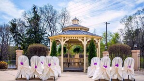 A gazebo with a dome roof, surrounded by chairs with white covers and purple accents, set on a paved area with trees and a fence in the background.