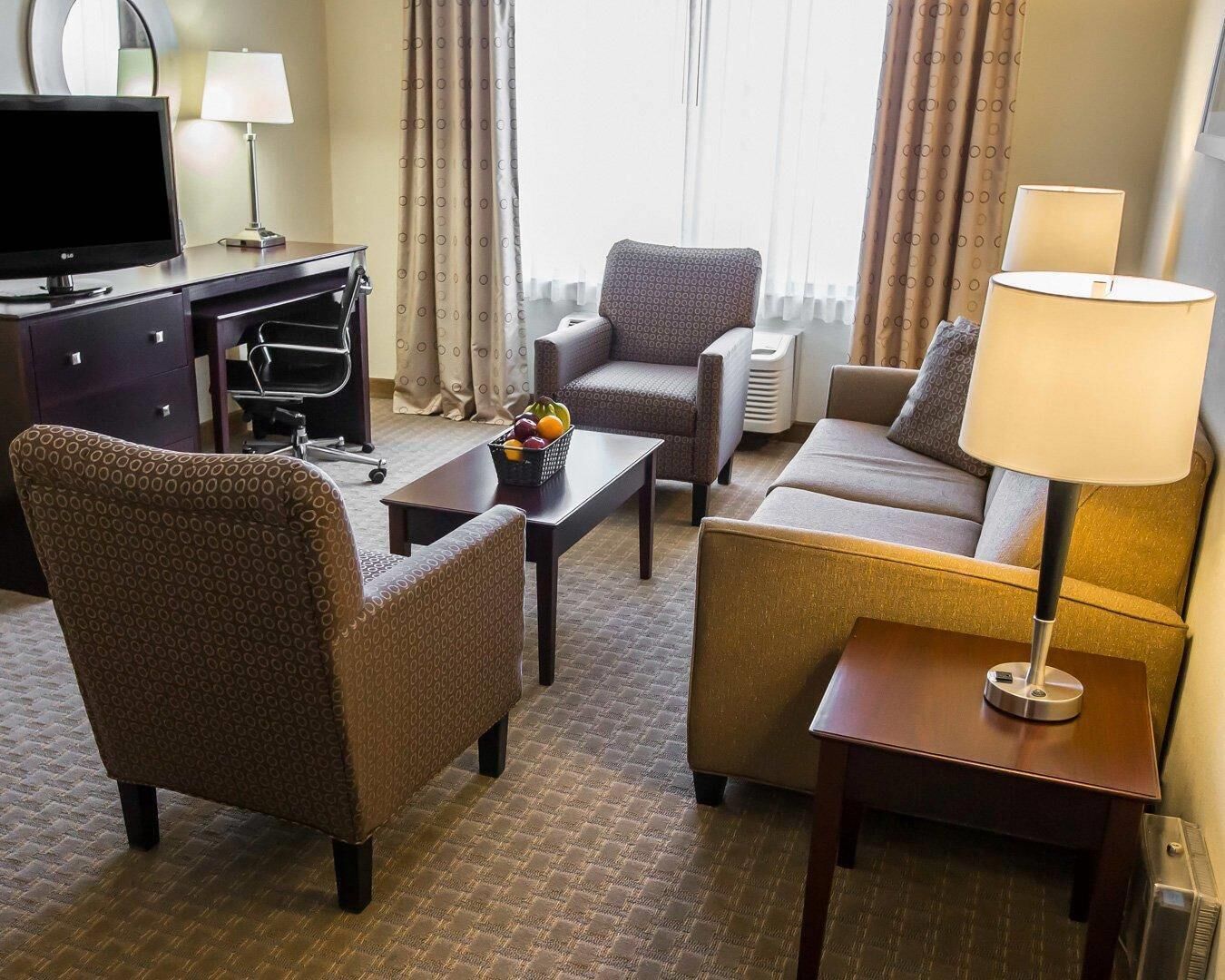 A hotel room with a sofa, armchair, desk, TV, lamp, and a fruit basket.