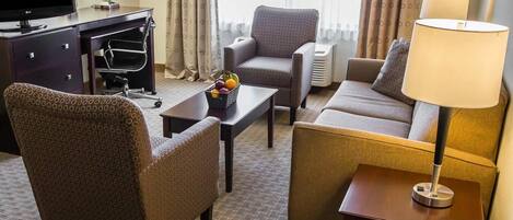 A hotel room with a sofa, armchair, desk, TV, lamp, and a fruit basket.