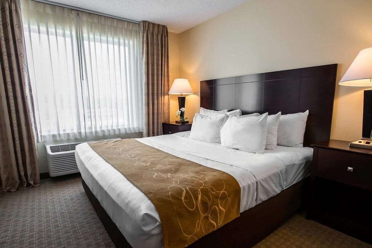 A hotel room with a large bed, two bedside lamps, a window with curtains, and a wall-mounted air conditioner.