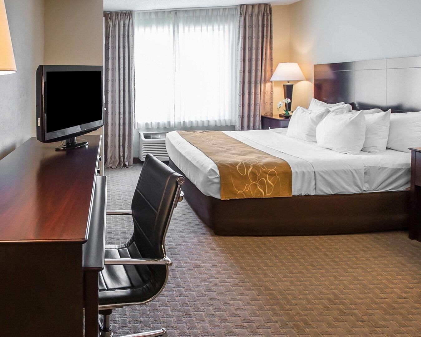 Una habitación de hotel con cama, escritorio, silla, televisor y ventana con cortinas.