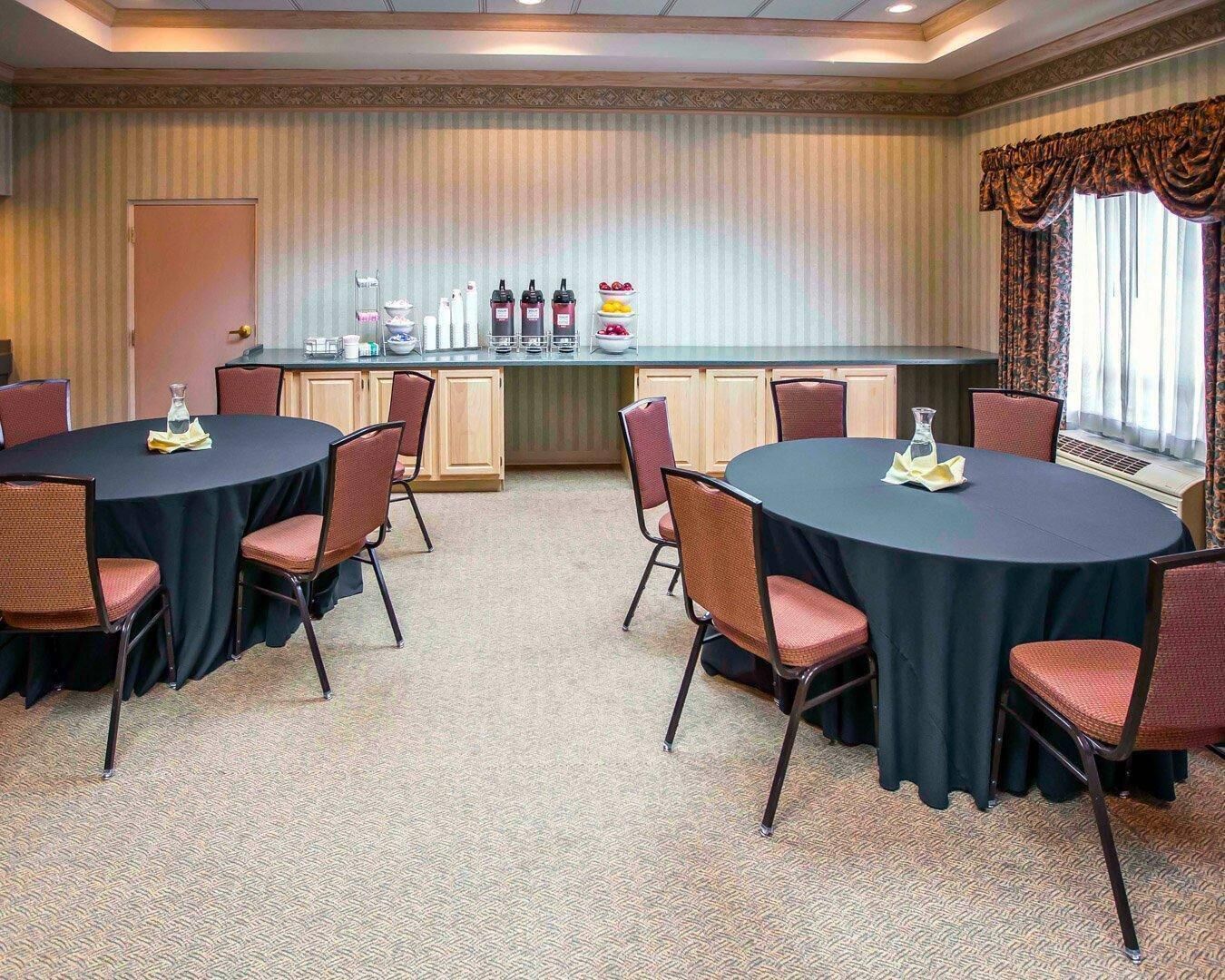Ein Konferenzraum mit zwei runden Tischen, Stühlen, einem Buffetbereich mit Beilagen und einem Fenster mit Vorhängen.