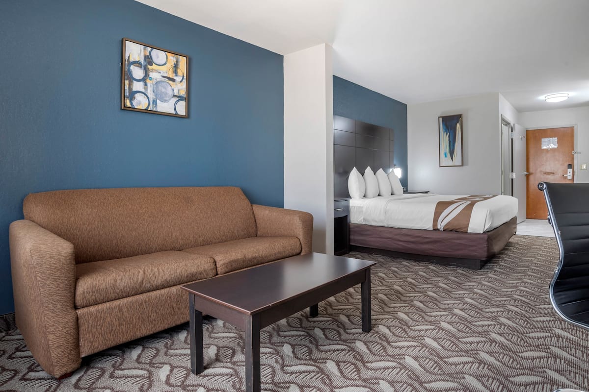 Habitación de hotel con un sofá marrón, una mesita pequeña, una cama con almohadas y una mesita de noche.