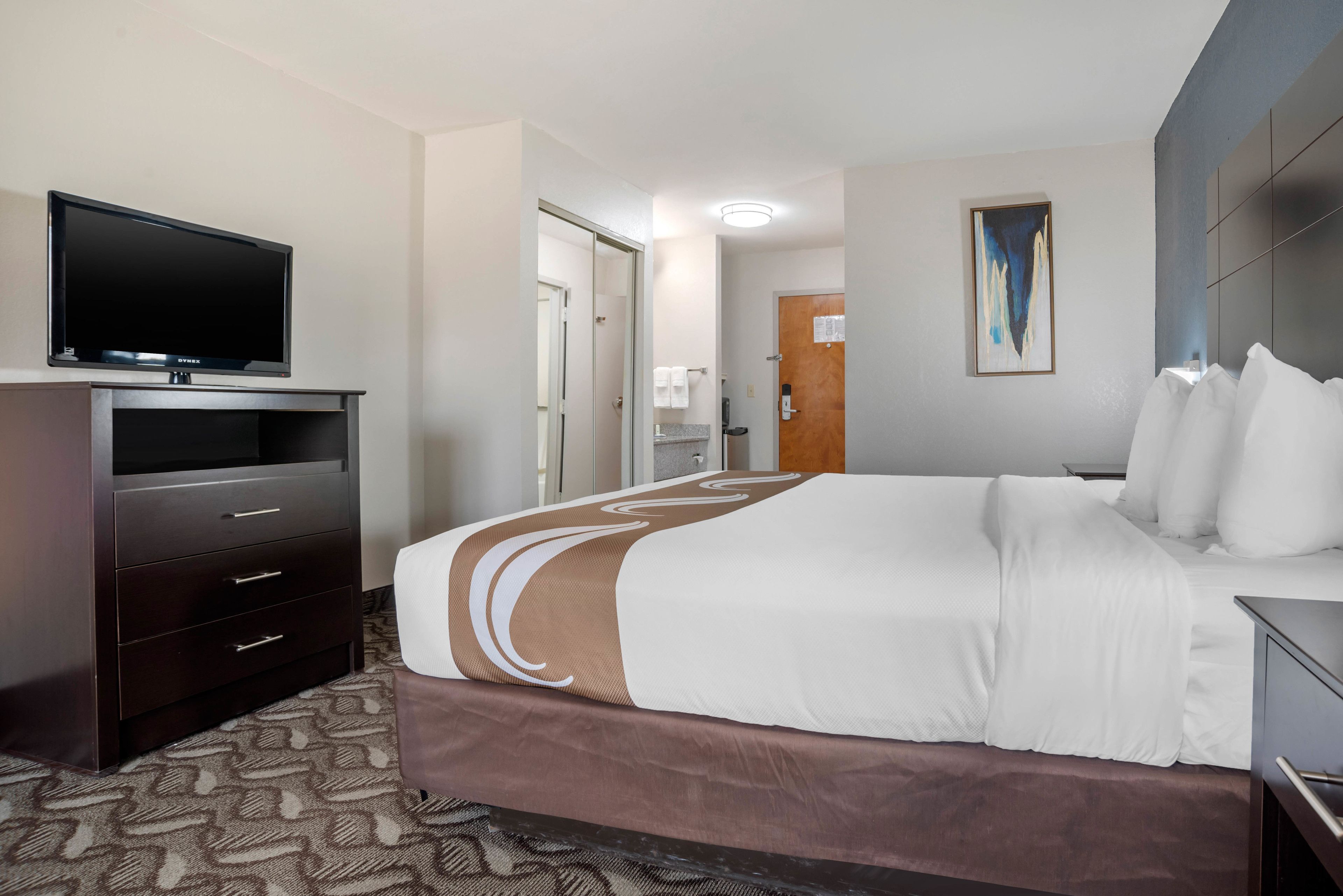 A hotel room with a bed, a TV on a stand, a dresser, and a painting on the wall.