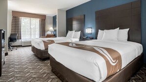 A hotel room with two beds, a desk, a chair, a lamp, and a window with curtains. - Quality Suites Baton Rouge East - Denham Springs (Baton Rouge)