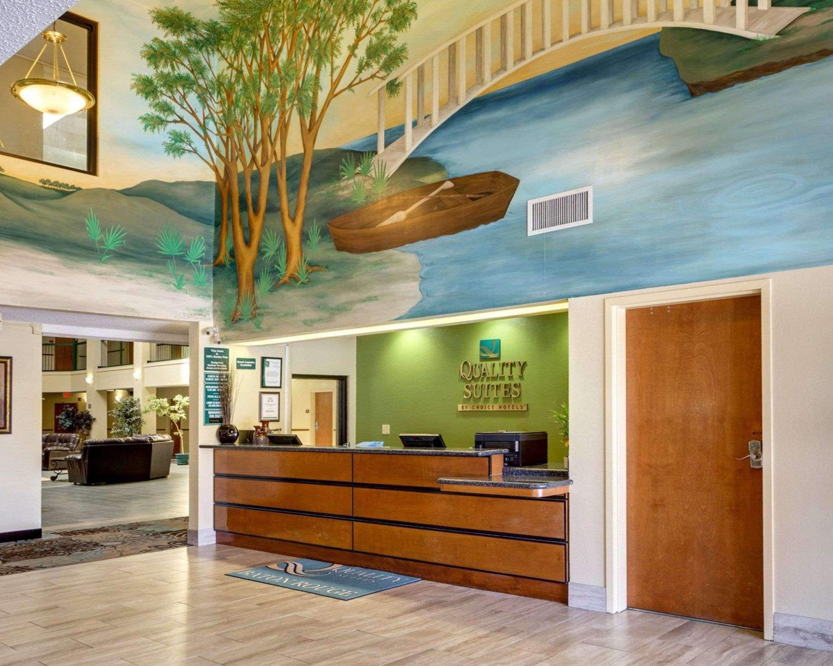 A hotel reception area with a mural of a river and trees, a reception desk, and a staircase.