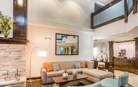 A modern hotel lobby with a stone fireplace, a large mirror, a sofa with patterned cushions, a coffee table, and a seating area with two armchairs.