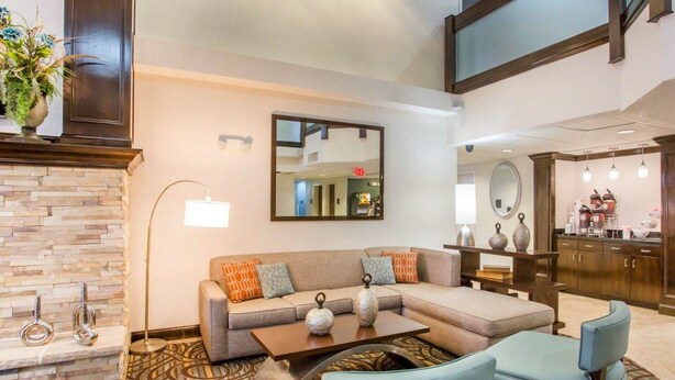 Un vestíbulo de hotel moderno con chimenea de piedra, un espejo grande, un sofá con cojines estampados, una mesa de centro y una zona de estar con dos sillones.