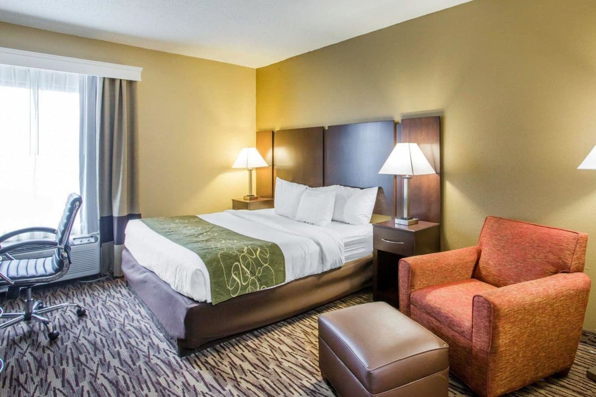 A hotel room with a bed, two bedside lamps, a chair, an ottoman, and a window with curtains.