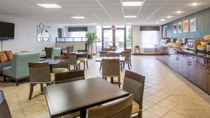 Una cafetería amplia con numerosas mesas y sillas, una barra grande con productos alimenticios, y un televisor montado en la pared.