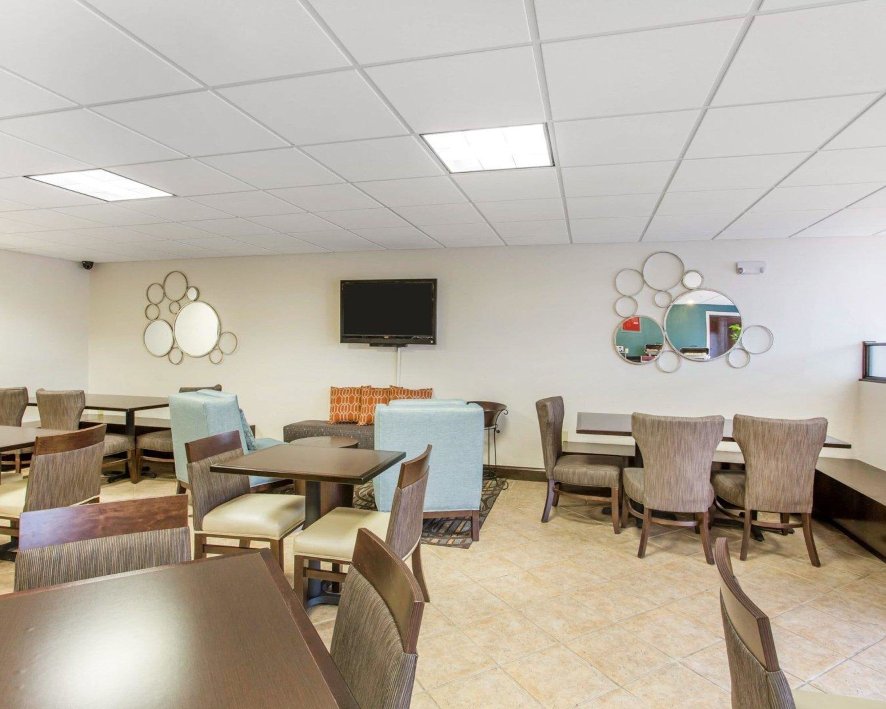 A hotel room with a dining area featuring multiple tables and chairs, a flat-screen TV mounted on the wall, and decorative wall mirrors.
