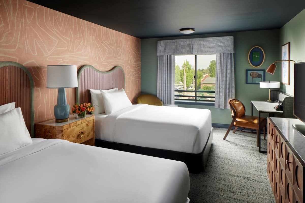 A hotel room with two beds, a desk, a chair, a TV, and a window with a view of the outside.