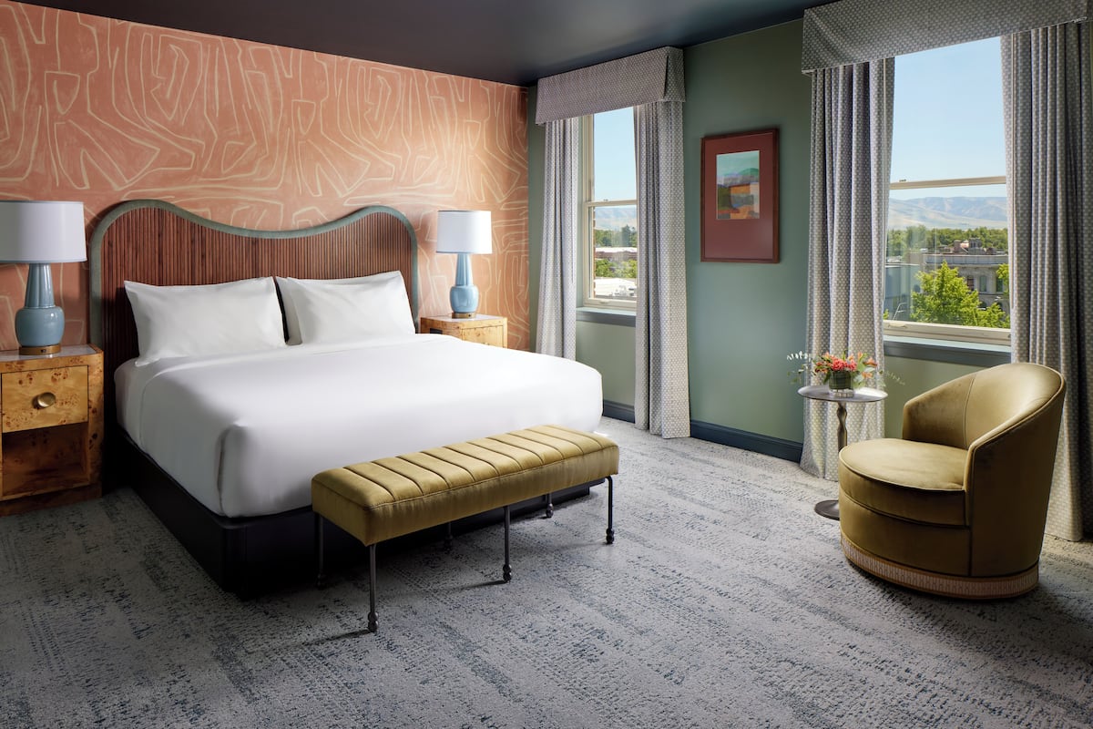 A hotel room with a large bed, two bedside tables with lamps, a bench, a chair, a small table with flowers, and a window with a view of the mountains.