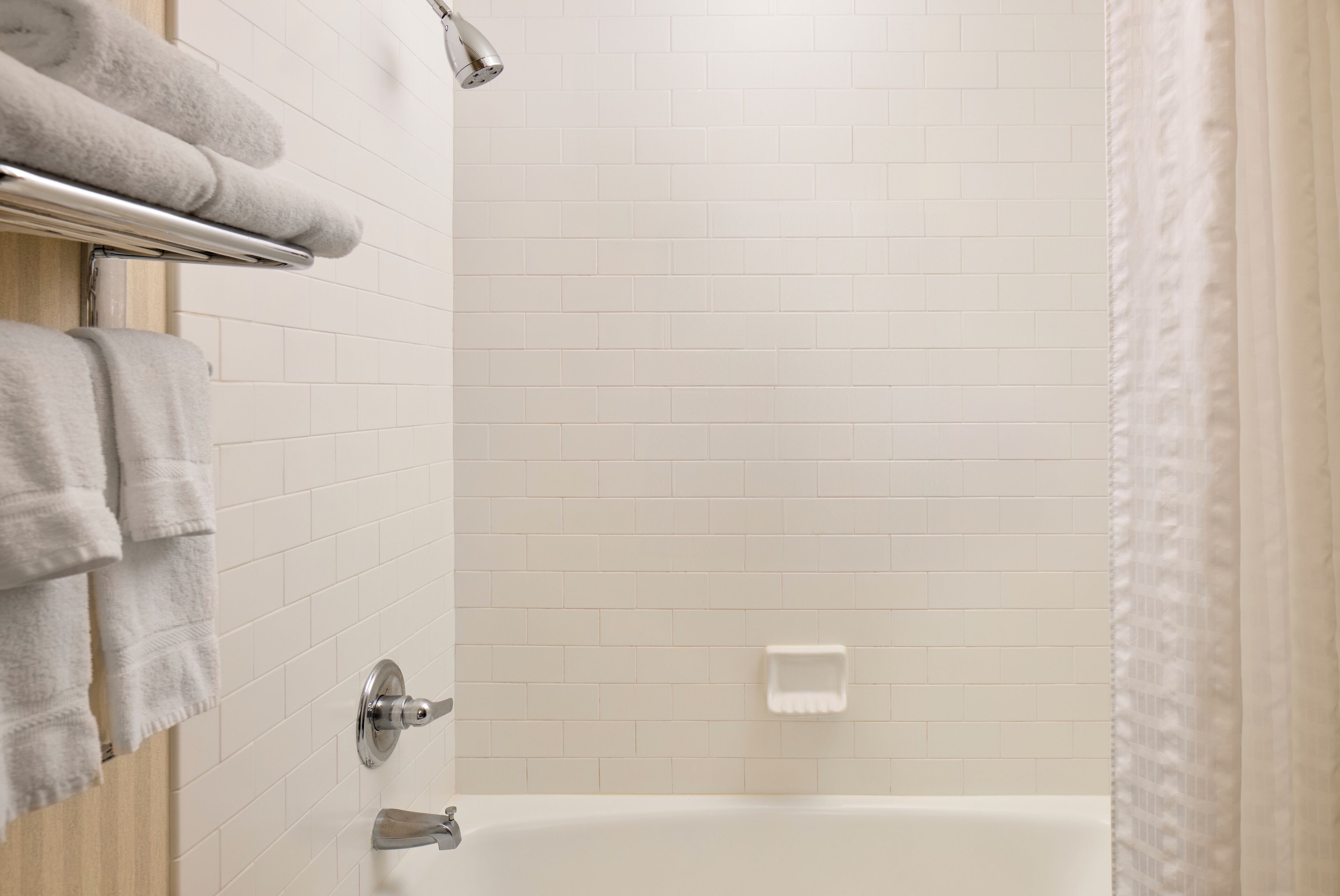 A bathroom with white tiled walls, a bathtub, a towel rack with folded towels, and a shower curtain.