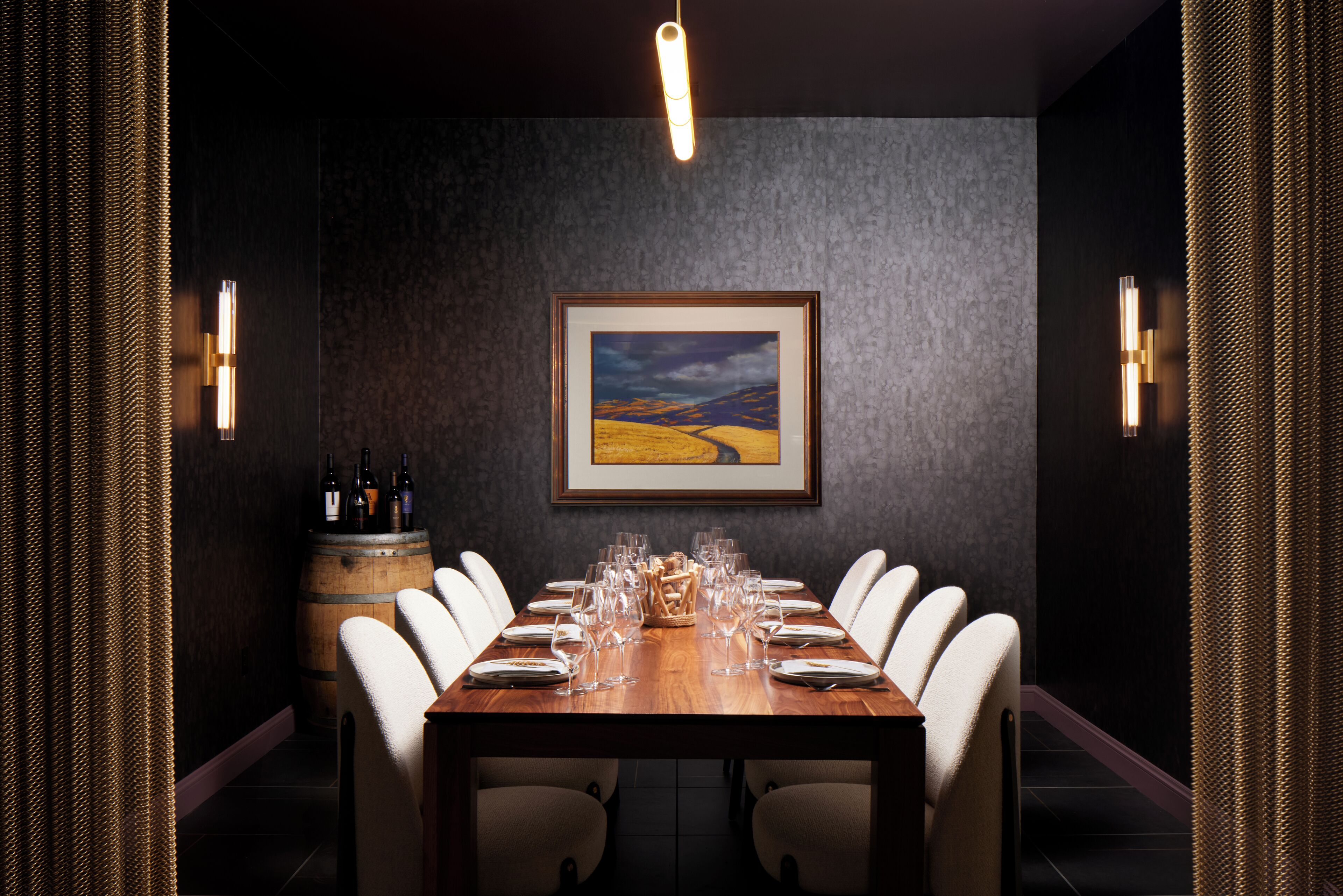 A dining area with a long wooden table set for six, surrounded by white chairs, a wooden barrel, and a framed painting on the wall.