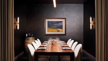 A dining area with a long wooden table set for six, surrounded by white chairs, a wooden barrel, and a framed painting on the wall.