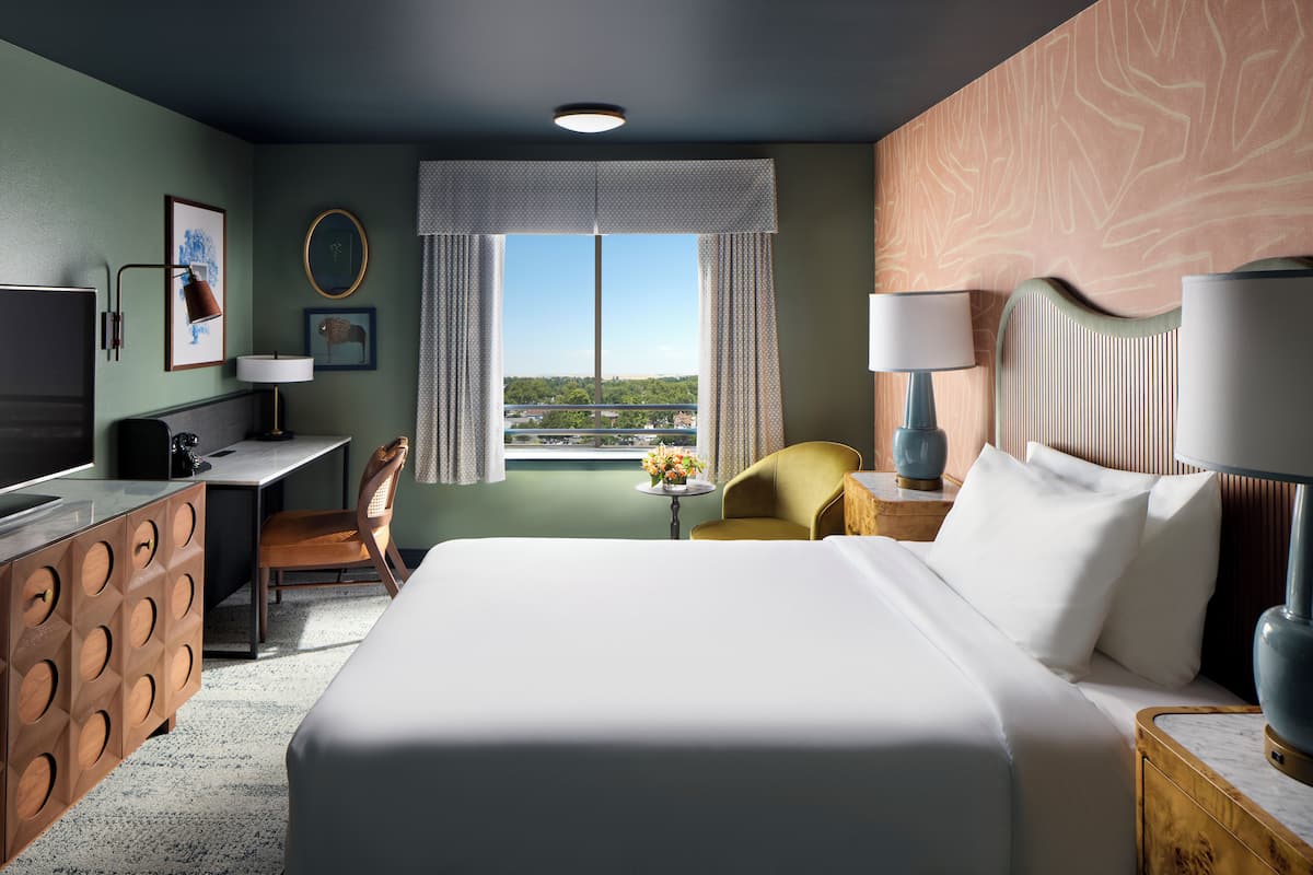 A hotel room with a large bed, a desk, a chair, a TV, and a view of the city through the window.