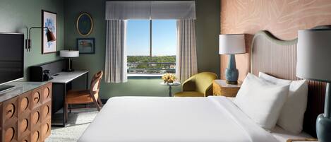 A hotel room with a large bed, a desk, a chair, a TV, and a view of the city through the window.