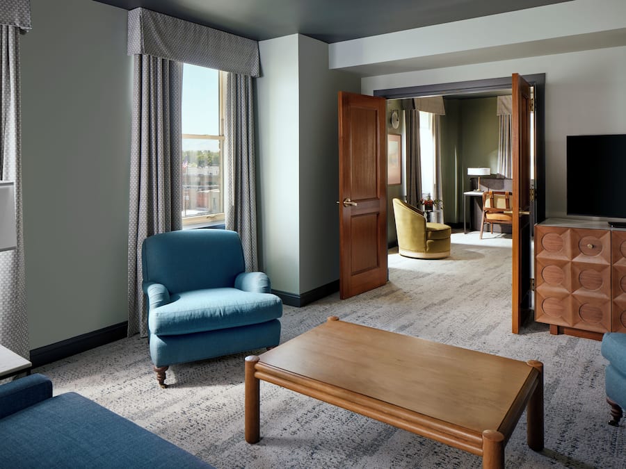 Habitación de hotel con un sillón azul, una mesa de centro de madera, un televisor y una puerta que da a otra habitación con un sillón amarillo y una lámpara.