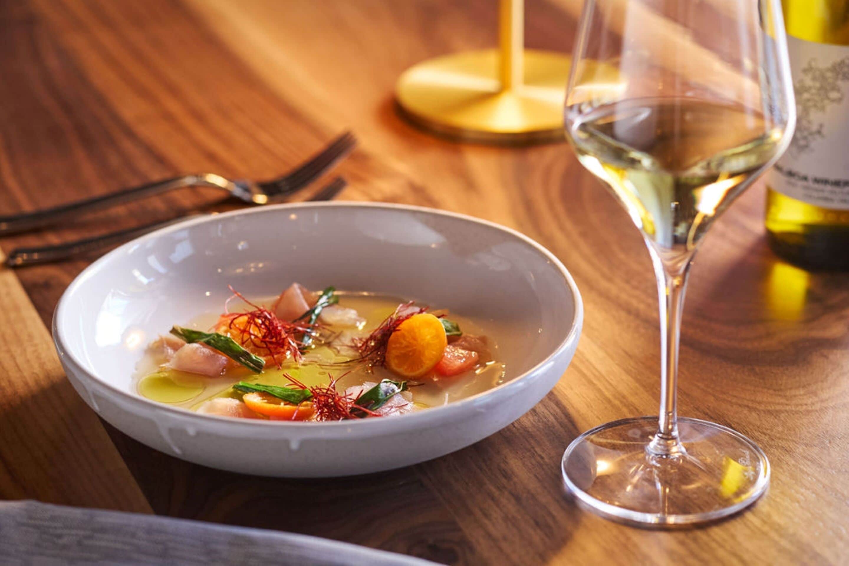 Un tazón de sopa con caldo claro, adornado con hierbas y verduras, y una copa de vino blanco sobre una mesa de madera.