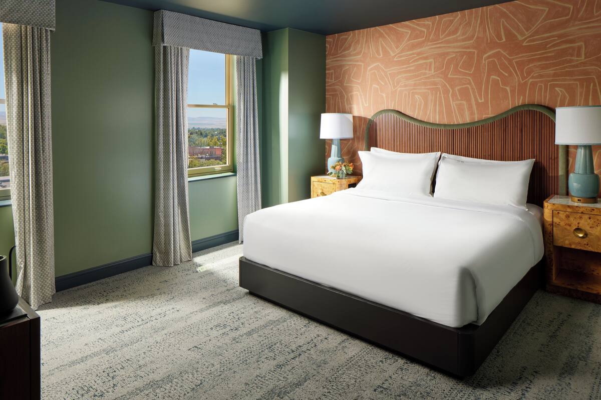 A hotel room with a large bed, two bedside tables with lamps, a view through a window, and a patterned wall.