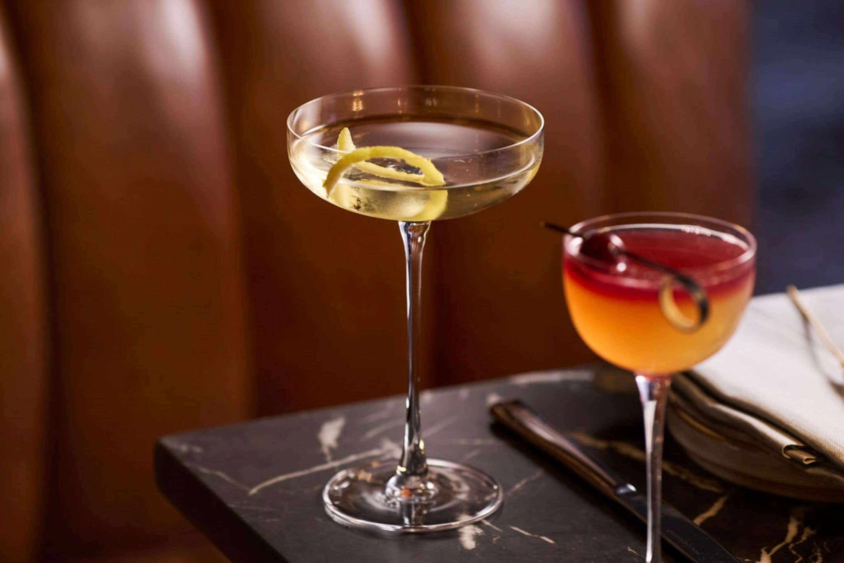 Two cocktails on a table with a napkin and cutlery beside them.