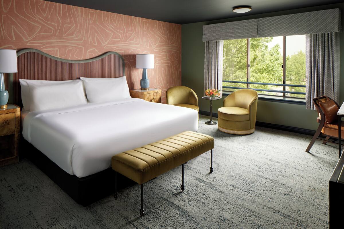 A hotel room with a large bed, a bench, two chairs, a nightstand with a lamp, and a window with a view of trees.