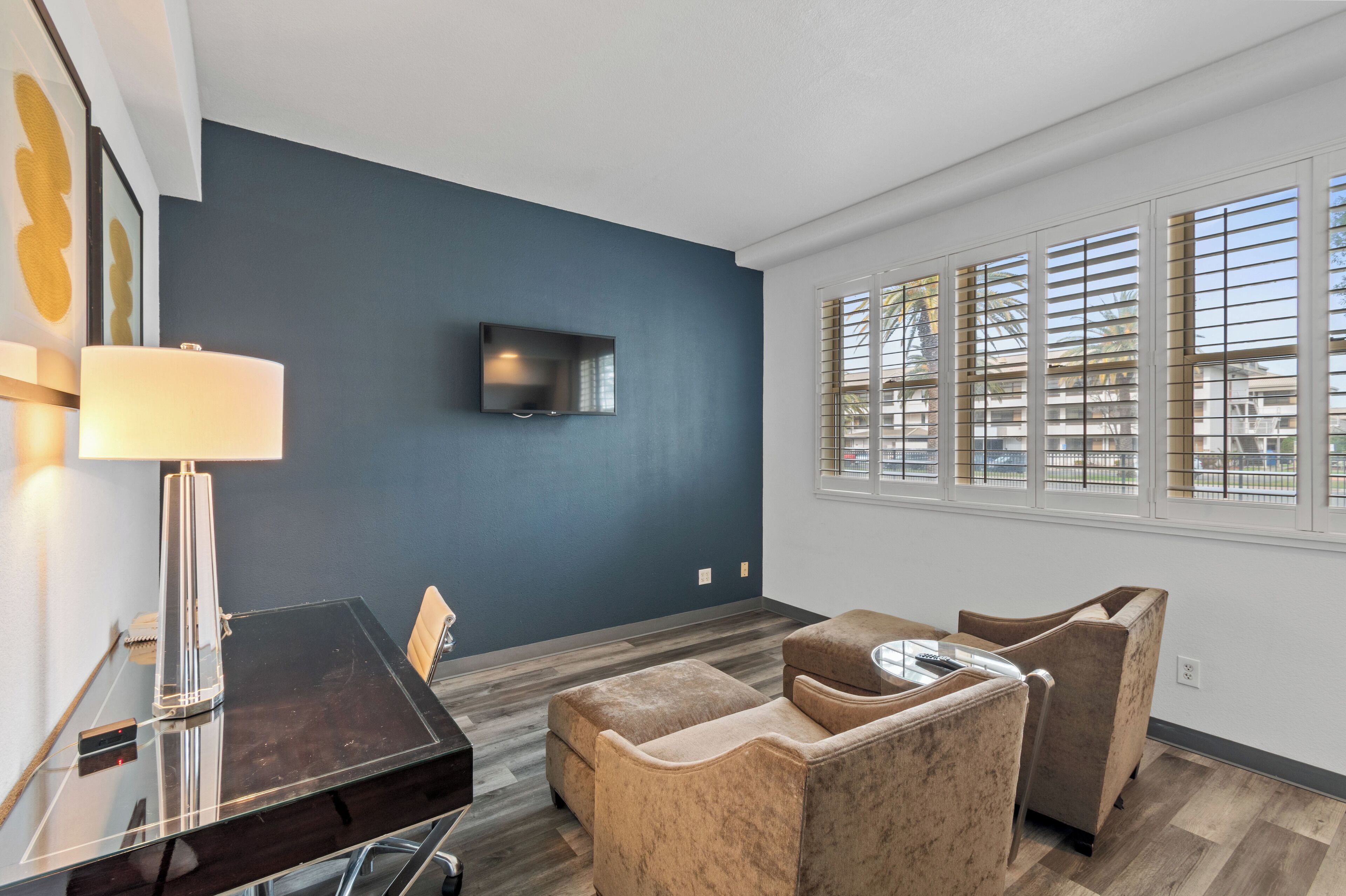 Un salon moderne avec un mur d’accent bleu foncé, un téléviseur à écran plat fixé au-dessus d’un parquet en bois foncé, et une grande fenêtre avec des stores.