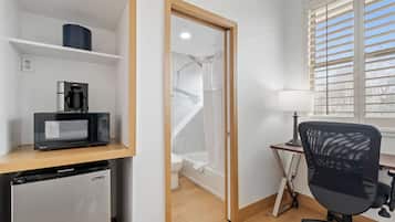 A compact room with a microwave, a small refrigerator, a desk with a chair, and a window with blinds.