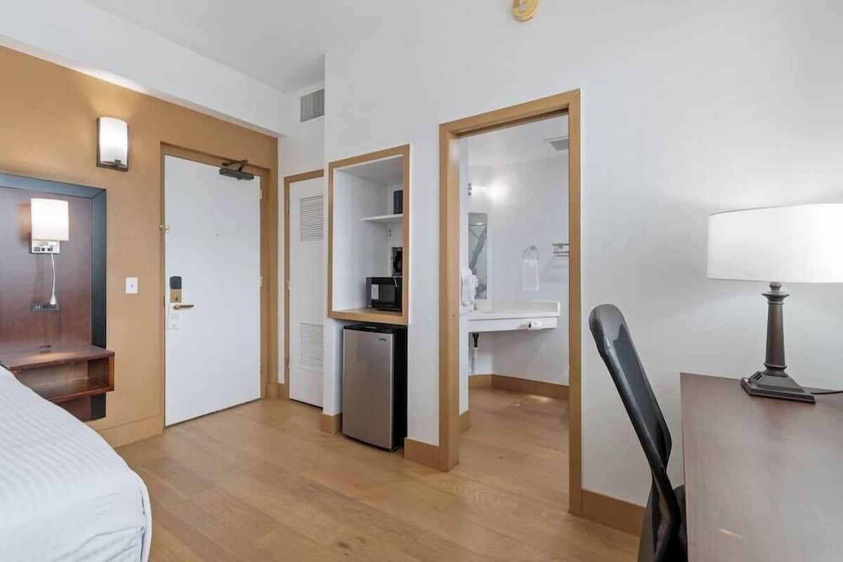 A hotel room with a bed, a desk, a chair, a refrigerator, and a microwave.