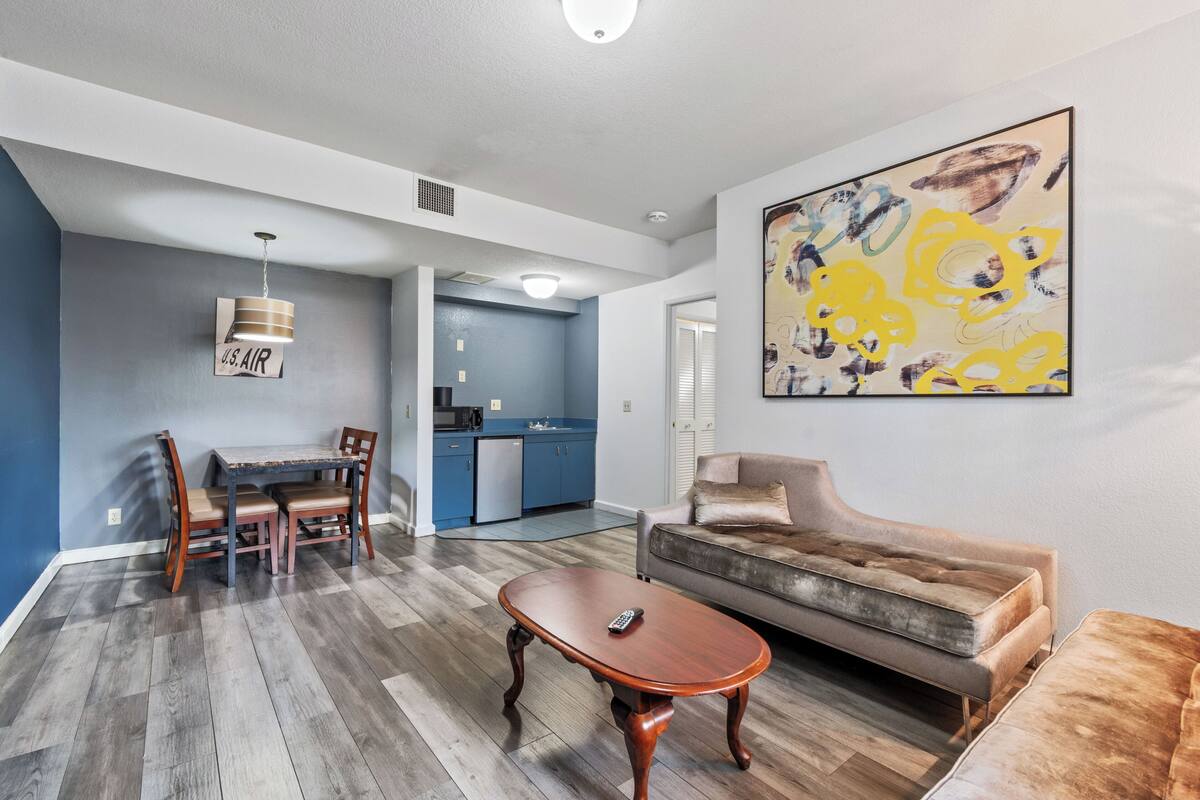 A modern living room with a dining area, a kitchen in the background, and a large abstract painting on the wall.