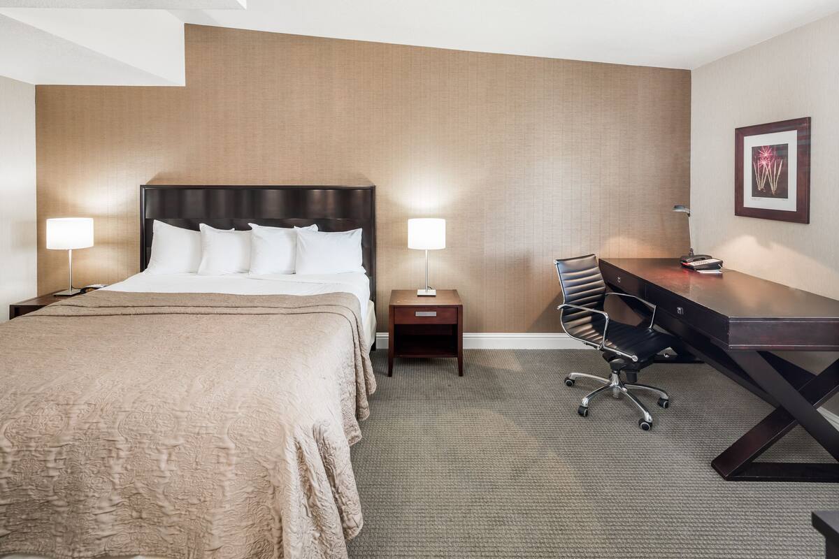 A hotel room with a large bed, a desk, and a chair. There is a nightstand with a lamp and a small table with a lamp.