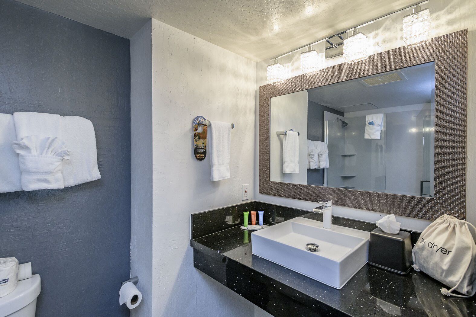 A modern bathroom with a black countertop, white sink, and a large mirror with a textured frame. There are towels hanging on the wall and a small towel rack.