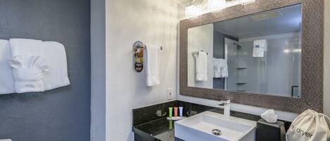 A modern bathroom with a black countertop, white sink, and a large mirror with a textured frame. There are towels hanging on the wall and a small towel rack.