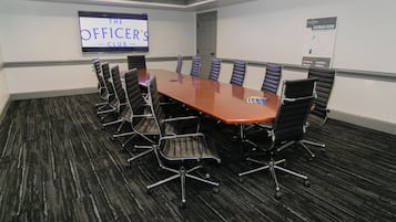 Een vergaderruimte met een lange tafel, zwarte stoelen, een projectiescherm en een vloerbedekking.