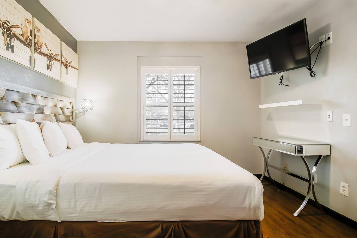 A bedroom with a bed, a window with blinds, a television mounted on the wall, and a desk with a chair.