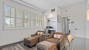 A modern living room with two brown armchairs, a small round table, and a sofa. There is a kitchen area with a microwave and a coffee maker visible.