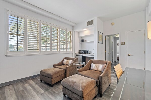 A modern living room with two brown armchairs, a small round table, and a sofa. There is a kitchen area with a microwave and a coffee maker visible. - Lions Gate Hotel (McClellan)