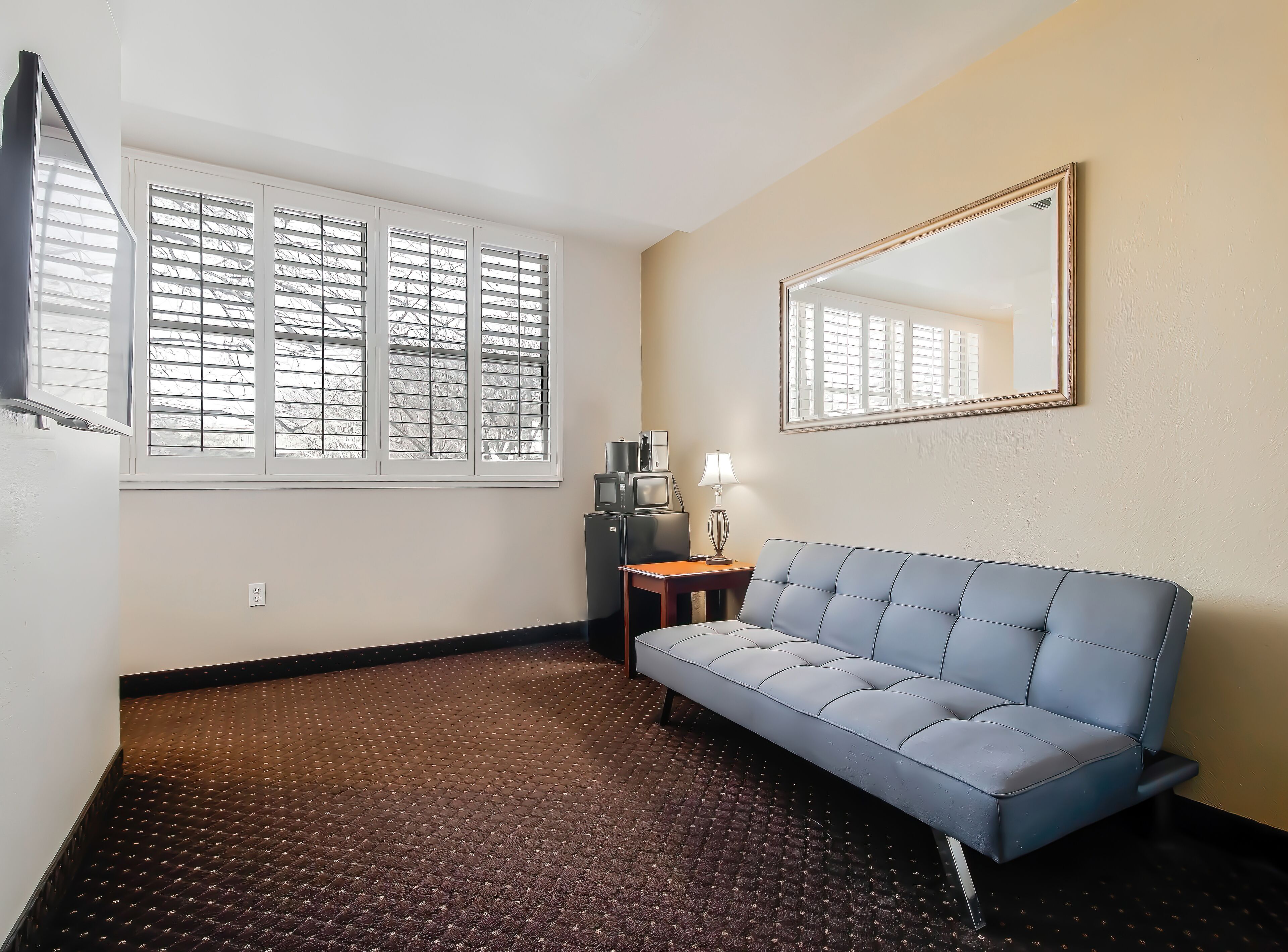 A room with a blue sofa, a small table with a lamp, a TV, and a window with blinds.