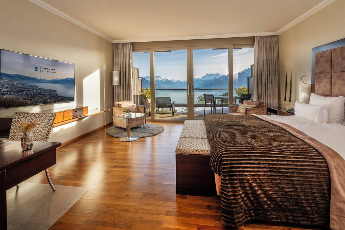 Ein Hotelzimmer mit einem großen Bett, einem Schreibtisch, einem Sessel, einer Couch, einem Fernseher und einem Balkon mit Bergblick.