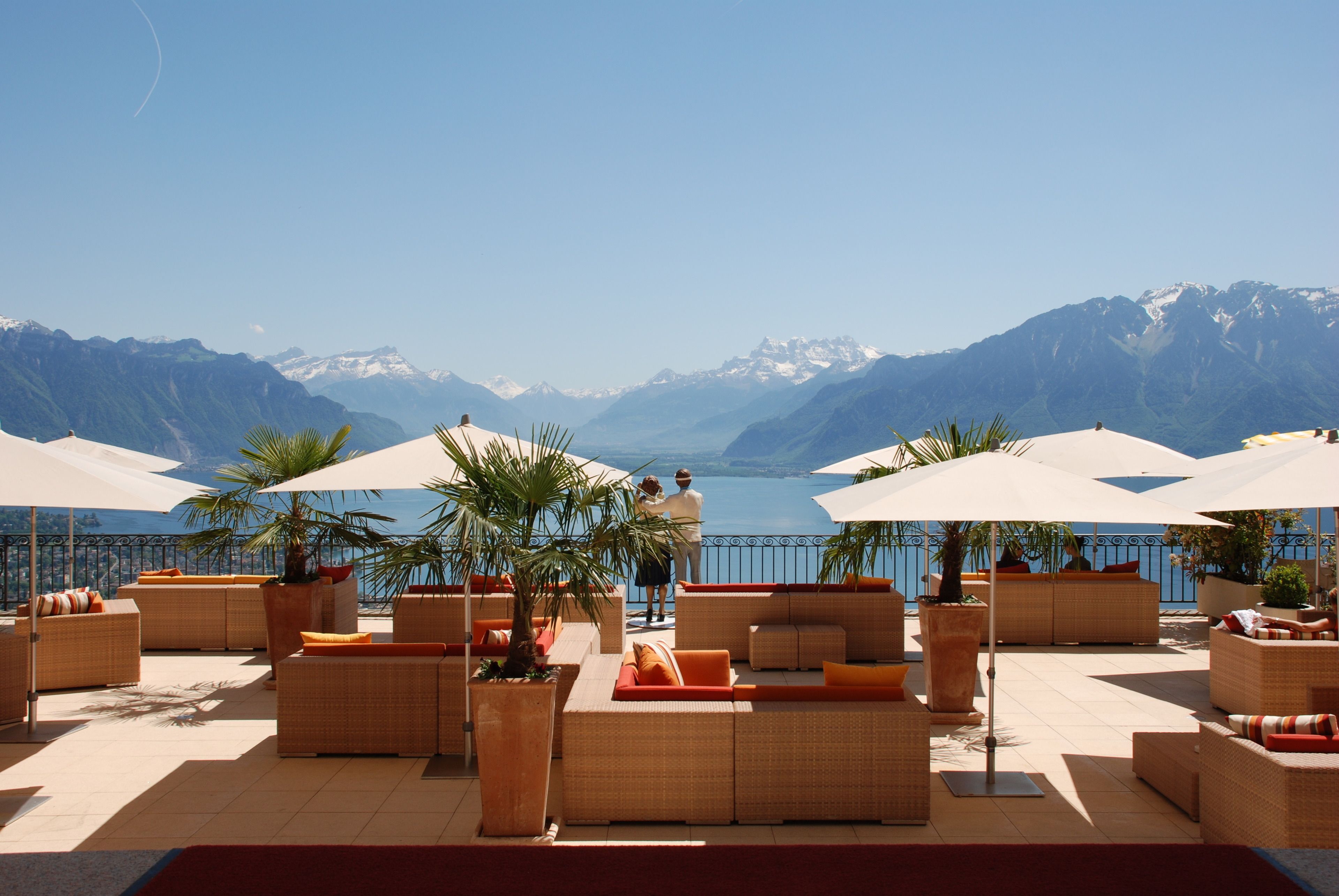 A terrace with wicker furniture and umbrellas, overlooking a lake and mountains.