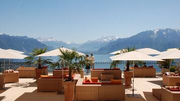 A terrace with wicker furniture and umbrellas, overlooking a lake and mountains.