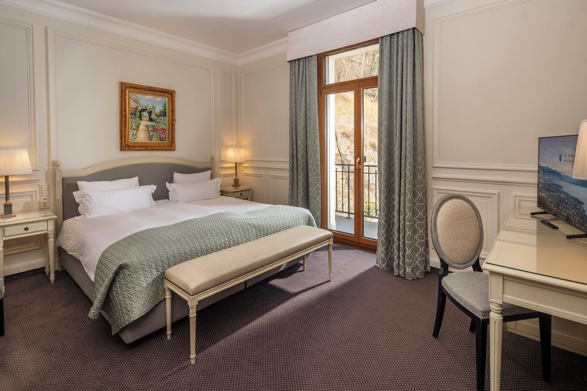 Ein Hotelzimmer mit Bett, Nachttischen, Schreibtisch, Stuhl, einem Fenster mit Vorhängen und einem Bild an der Wand.