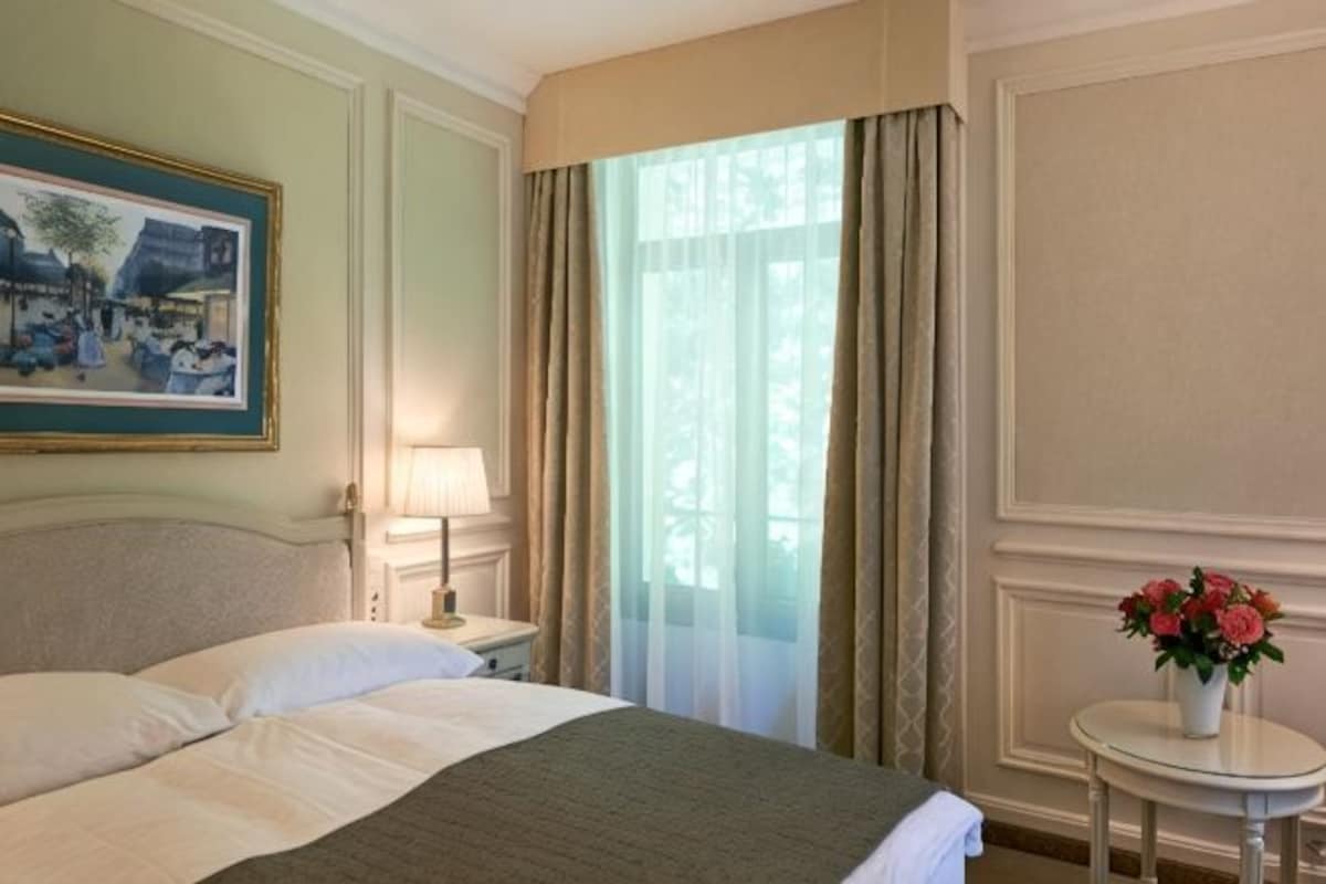 Habitación de hotel con cama, mesita de noche con lámpara, ventana con cortinas y un cuadro en la pared.