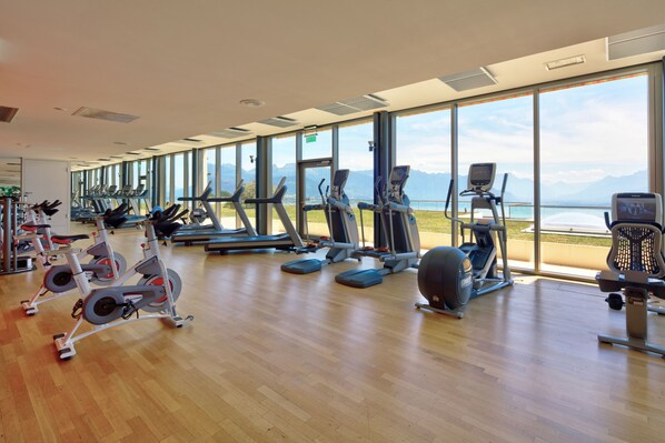 Un gimnasio con diversas máquinas de ejercicio, ventanas amplias y vistas a la montaña.