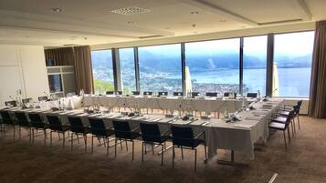 A conference room with a long table, chairs, microphones, and water bottles. Large windows offer a view of a city and a body of water.